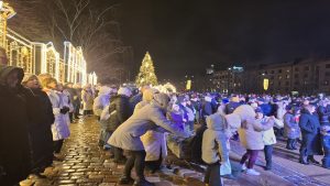 Cilvēku pūlis svin Jaungada sagaidīšanu pilsētas centrā ar izgaismotām ēkām un Ziemassvētku egli.