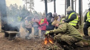 Militārais instruktors pie ugunskura mežā māca bērnu grupai prasmes ārpus telpām.