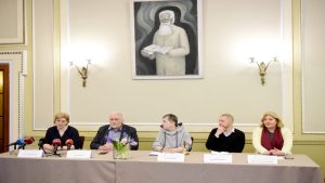 Preses konference ar pieciem dalībniekiem pie galda dekorēta ar ziediem un gleznu fonā.