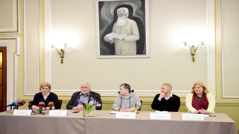 Preses konference ar pieciem dalībniekiem pie galda dekorēta ar ziediem un gleznu fonā.