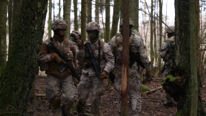 Kadeti militārajās apmācībās mežā, tērpti uniformās ar ieročiem rokās.