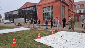 Skolēni piedalās izzinošā barikāžu laika aktivitātē pie sarkana vēsturiska ēkas fasādes Kuldīgā.