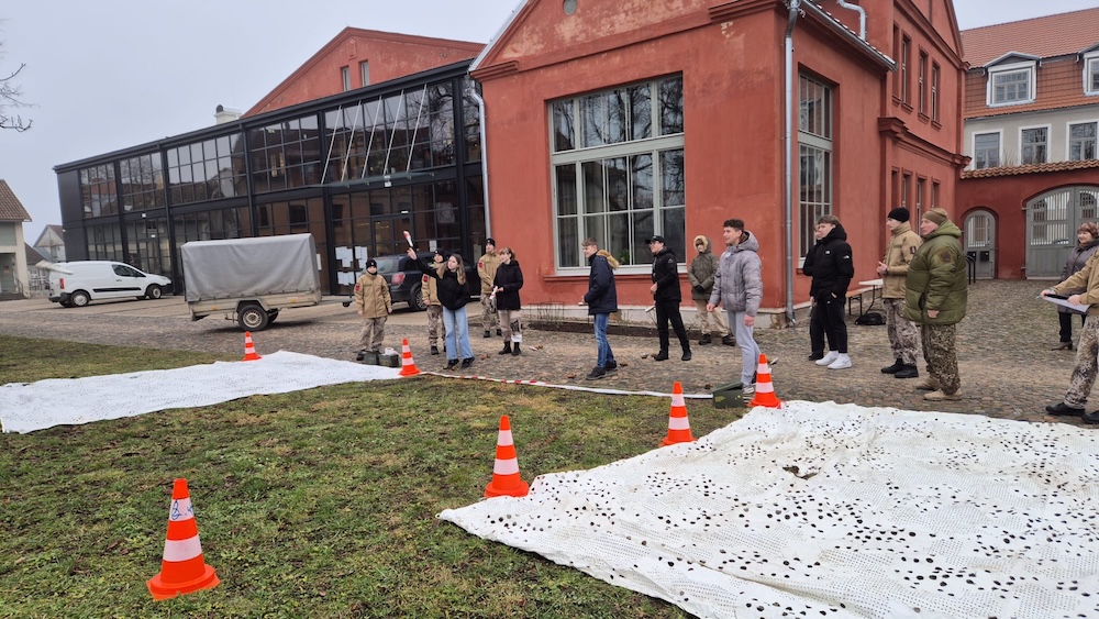 Skolēni piedalās izzinošā barikāžu laika aktivitātē pie sarkana vēsturiska ēkas fasādes Kuldīgā.