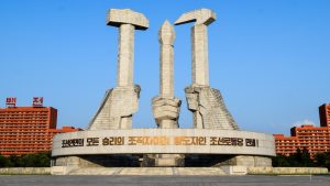 Monumentāla statuja Ziemeļkorejā ar trīs rokām, kas tur āmuru, sirpi un spalvu