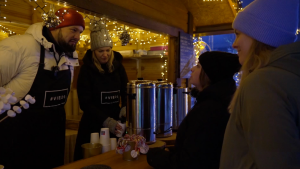 Divi cilvēki ar melniem priekšautiem, kas apkalpo klientus ziemas tirdziņā, apkārt ir Ziemassvētku rotājum