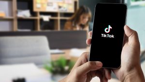 Cilvēks tur rokās viedtālruni ar atvērtu TikTok aplikāciju, redzama TikTok logo melnā ekrānā.