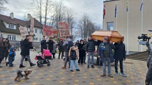 Protestētāji pie pašvaldības ēkas, nesot simbolisku zārku un plakātus ar uzrakstiem.