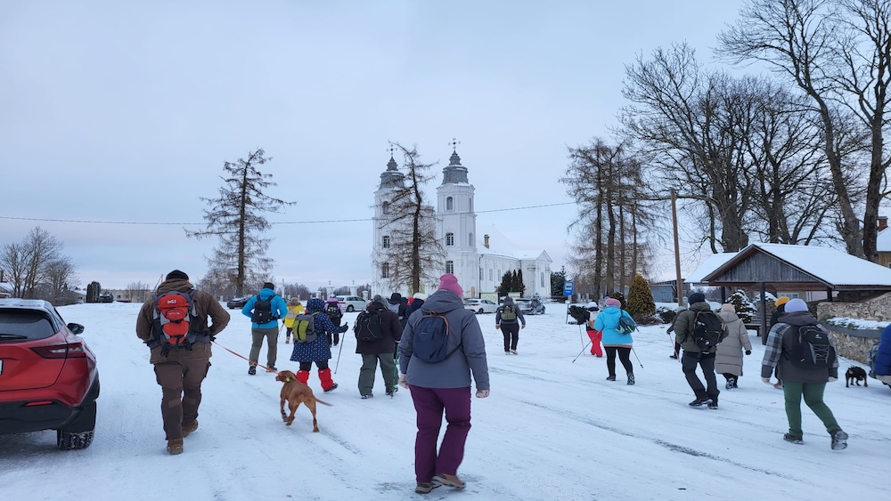 Cilvēki ziemas pārgājienā dodas uz baznīcu, kas redzama fonā.