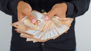 Rokas tur kaudzīti ar 50 eiro banknotēm, simbolizējot naudu un finanšu darījumus
