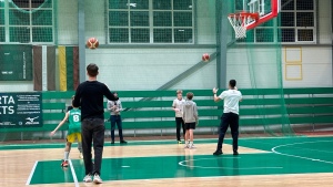 Basketbola nodarbība sporta zālē, treneris un jaunieši trenē metienus grozā.