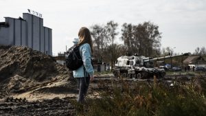 Jauna sieviete ar mugursomu stāv kara postītā teritorijā, kur redzams tanks un sabojātas ēkas.