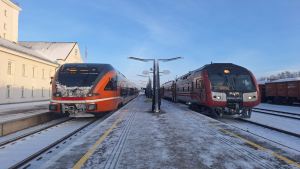 Divi vilcieni stacijā ziemas laikā, viens oranžs elektriskais un otrs sarkans dīzeļvilciens.