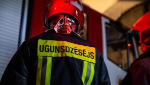Ugunsdzēsējs darba formā un ķiverē pie ugunsdzēsēju transporta.