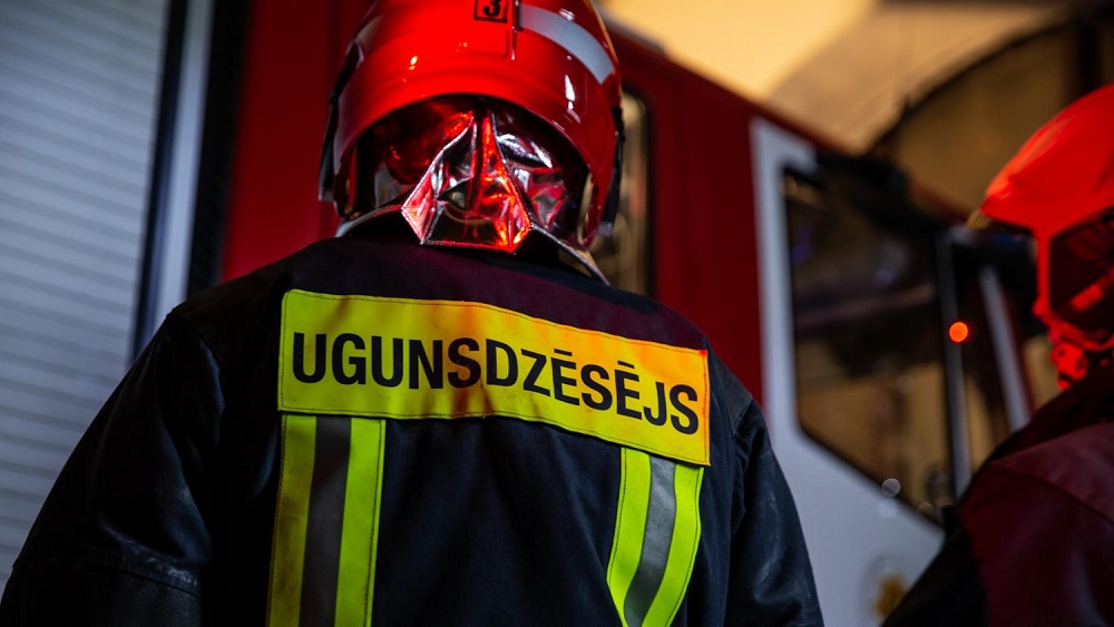 Ugunsdzēsējs darba formā un ķiverē pie ugunsdzēsēju transporta.