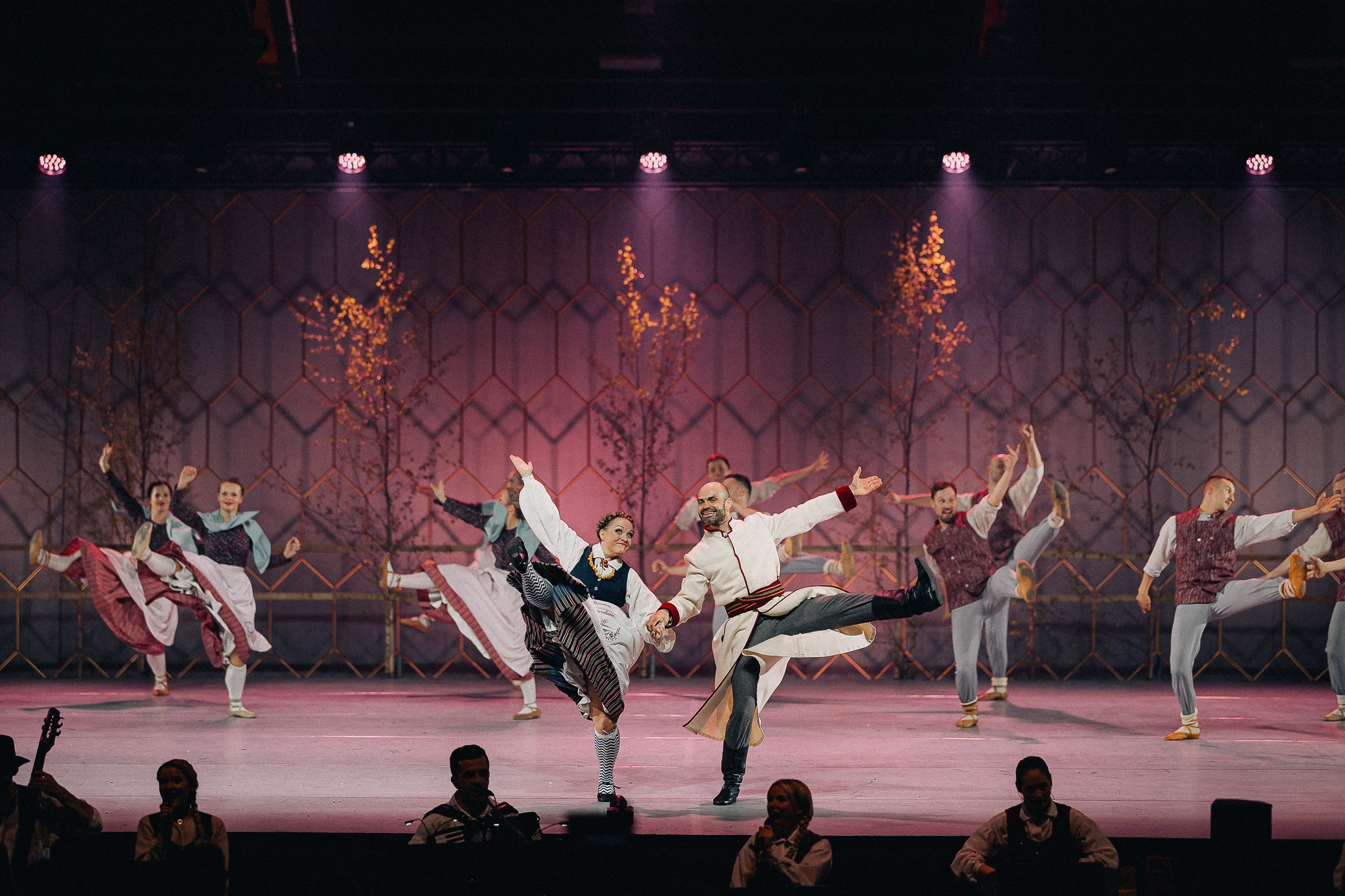 Latviešu tautas deju kolektīvs krāšņā uzstāšanās brīdī uz skatuves, dejotāji tradicionālajos tautastērpos.