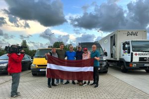 Cilvēku grupa tur Latvijas karogu humānās palīdzības transporta priekšā.