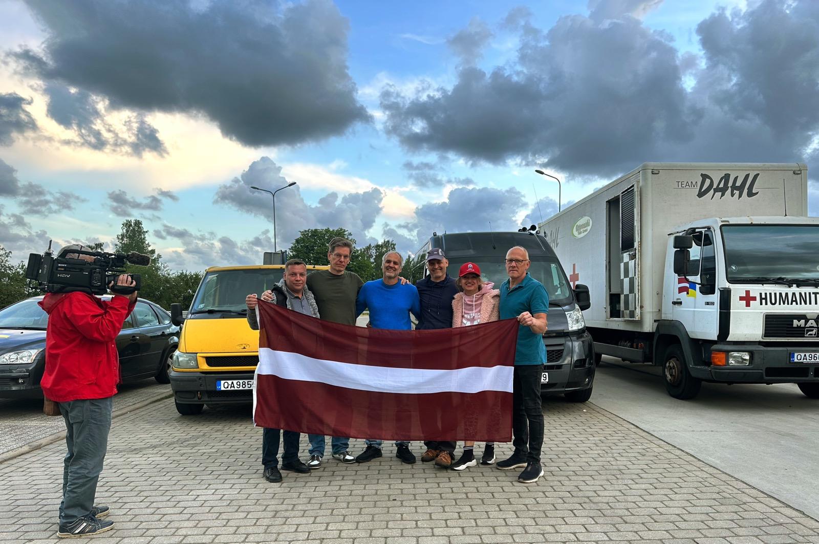 Cilvēku grupa tur Latvijas karogu humānās palīdzības transporta priekšā.