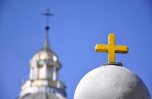 Dzeltenais krusts uz baznīcas kupola ar neskaidru baznīcas torni fonā, zilas debesis.