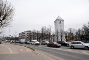 Satiksme pilsētā ar vēsturisku balto torni un daudzstāvu ēkām fonā.