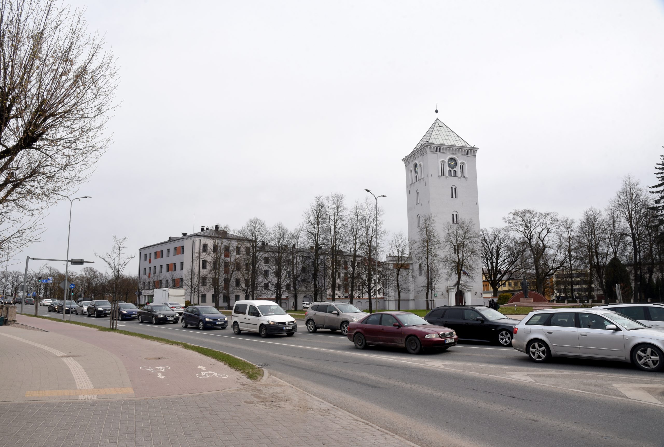 Satiksme pilsētā ar vēsturisku balto torni un daudzstāvu ēkām fonā.