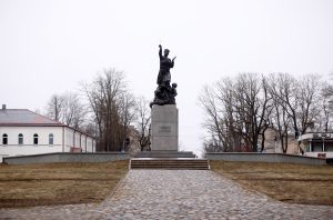 Piemineklis "Vienoti Latvijai" Rēzeknē uz bruģēta ceļa fona, kokiem bez lapām.
