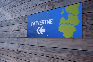 Zila patvertnes norādes plāksne ar Baltijas kartes siluetu, piestiprināta pie koka sienas.