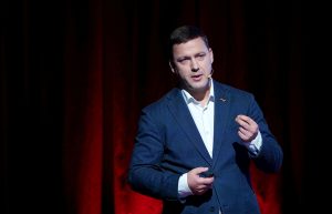 Vīrietis tumšā uzvalkā uzstājas uz skatuves ar mikrofonu pie auss, fonā sarkans aizkars