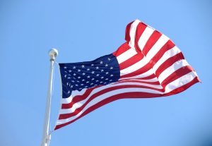 Amerikas Savienoto Valstu karogs plīvo vējā uz fona ar zilām debesīm.