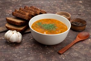 Bļoda ar dārzeņu zupu, rudzu maize, ķiploks un garšvielas uz koka galda.