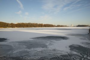 Daļēji aizsalis ezers ar sniega un ledus slāņiem ziemas ainavā.