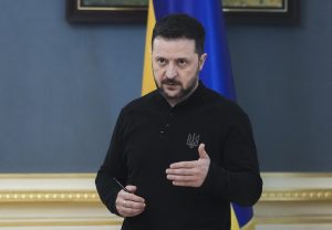 Volodimirs Zelenskis melnā kreklā ar valsts simbolu, runājot oficiālā pasākumā, fonā redzams zili-dzeltens karogs