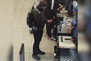 Vīrietis tumšā apģērbā ar mugursomu pie veikala pašapkalpošanās kases