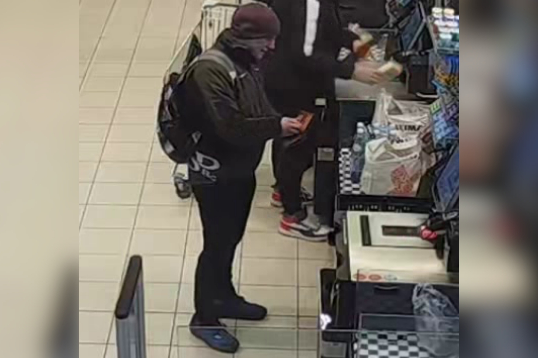 Vīrietis tumšā apģērbā ar mugursomu pie veikala pašapkalpošanās kases