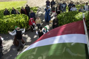 Sarkanbalti zaļš karogs priekšplānā, fonā tautas deju dejotāji un skatītāji pulcējas brīvdabas pasākumā