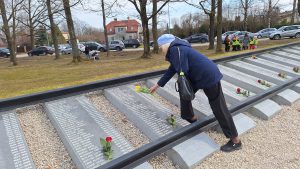 Sieviete zilā jakā un beretē noliek ziedus pie piemiņas plāksnes ar uzvārdiem.