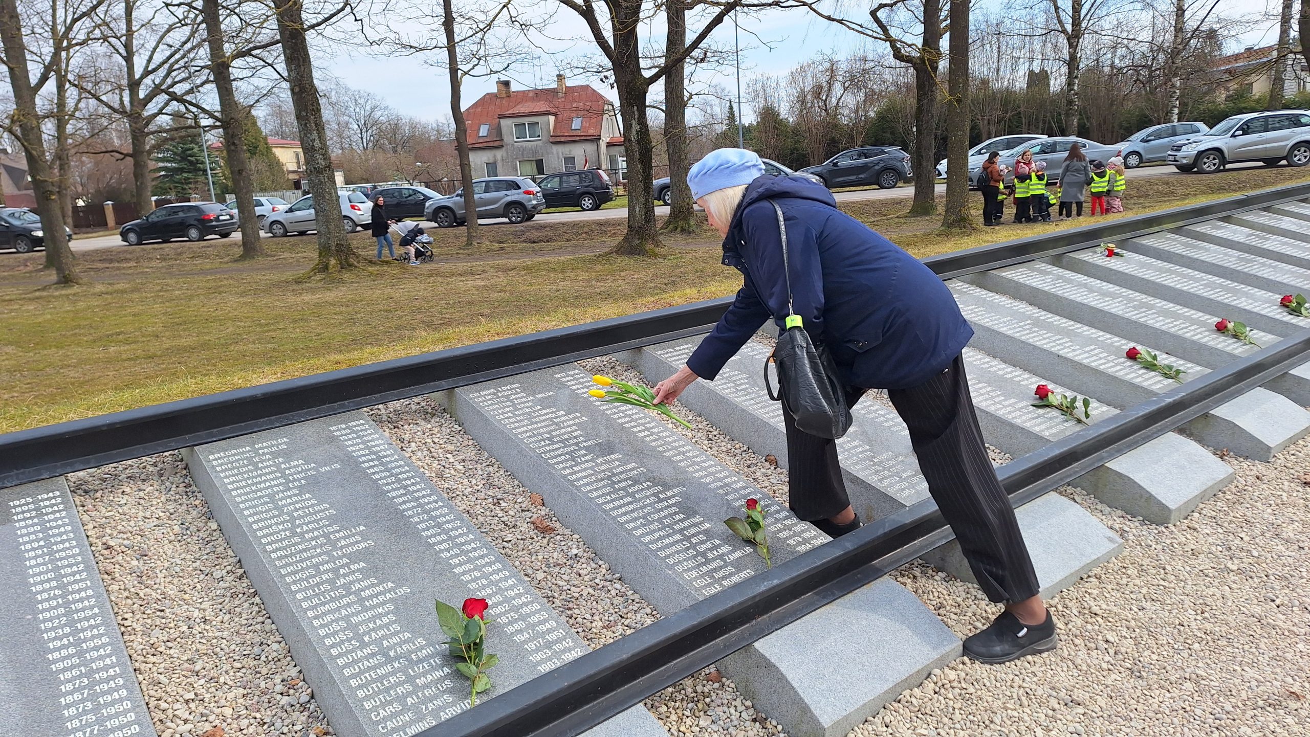 Sieviete zilā jakā un beretē noliek ziedus pie piemiņas plāksnes ar uzvārdiem.