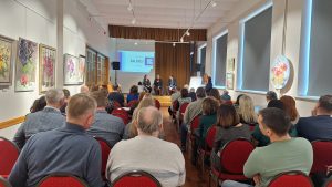 : Skats uz pasākumu ar cilvēkiem, kas piedalās diskusijā, un mākslas darbiem uz sienas.