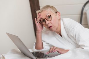 Sieviete ar īsiem, gaišiem matiem un brillēm, ģērbusies halātā, izskatās noraizējusies, skatoties datorā