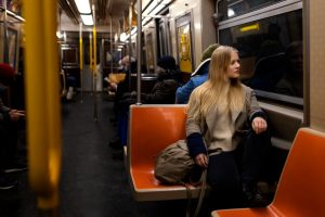 Sieviete ar gaišiem matiem, siltā apģērbā, sēž oranžā metro sēdeklī un skatās pa logu