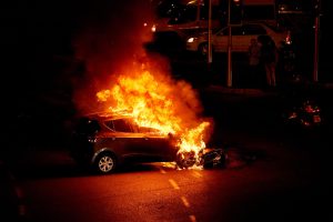 Degošs automobilis naktī ielas krustojumā ar liesmām, kas pārņēmušas motora daļu