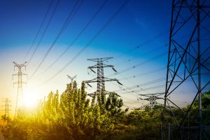 Augstsprieguma elektrolīniju torņi un vadi, kas slejas virs koku galotnēm saulrietā