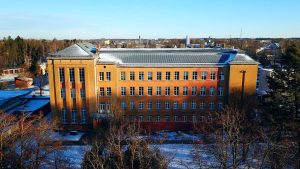 Vairāku stāvu ķieģeļu ēka ar sniega klātu jumtu, fonā redzama pilsētas ainava