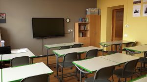 Skolas klase ar galdiem, krēsliem un interaktīvo ekrānu.