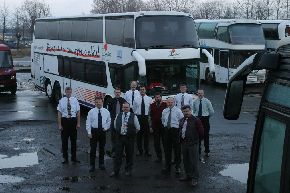 Grupas fotogrāfija ar autobusu vadītājiem, stāvot pie divstāvu tūrisma autobusa stāvlaukumā.