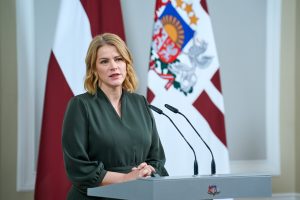 Premjere Evika Siliņa zaļā kleitā runā preses konferencē, fonā redzami Latvijas karogi un valsts ģerbonis