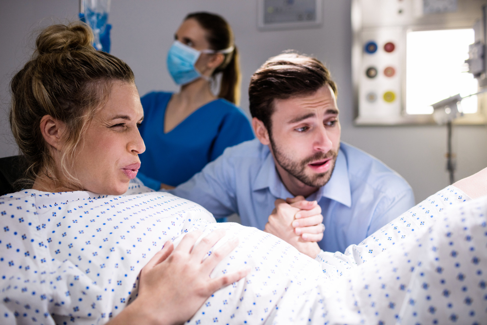 Sieviete dzemdību procesā, vīrietis tur viņas roku, fonā mediķe ar masku