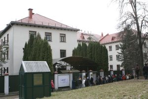 Cilvēku rinda pie liela, gaiša ēku kompleksa ar sarkanu jumtu un augstiem kokiem priekšplānā