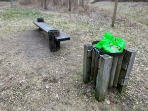 Koka atkritumu tvertne ar zaļu plastmasas maisu un blakus esošu soliņu dabas teritorijā.