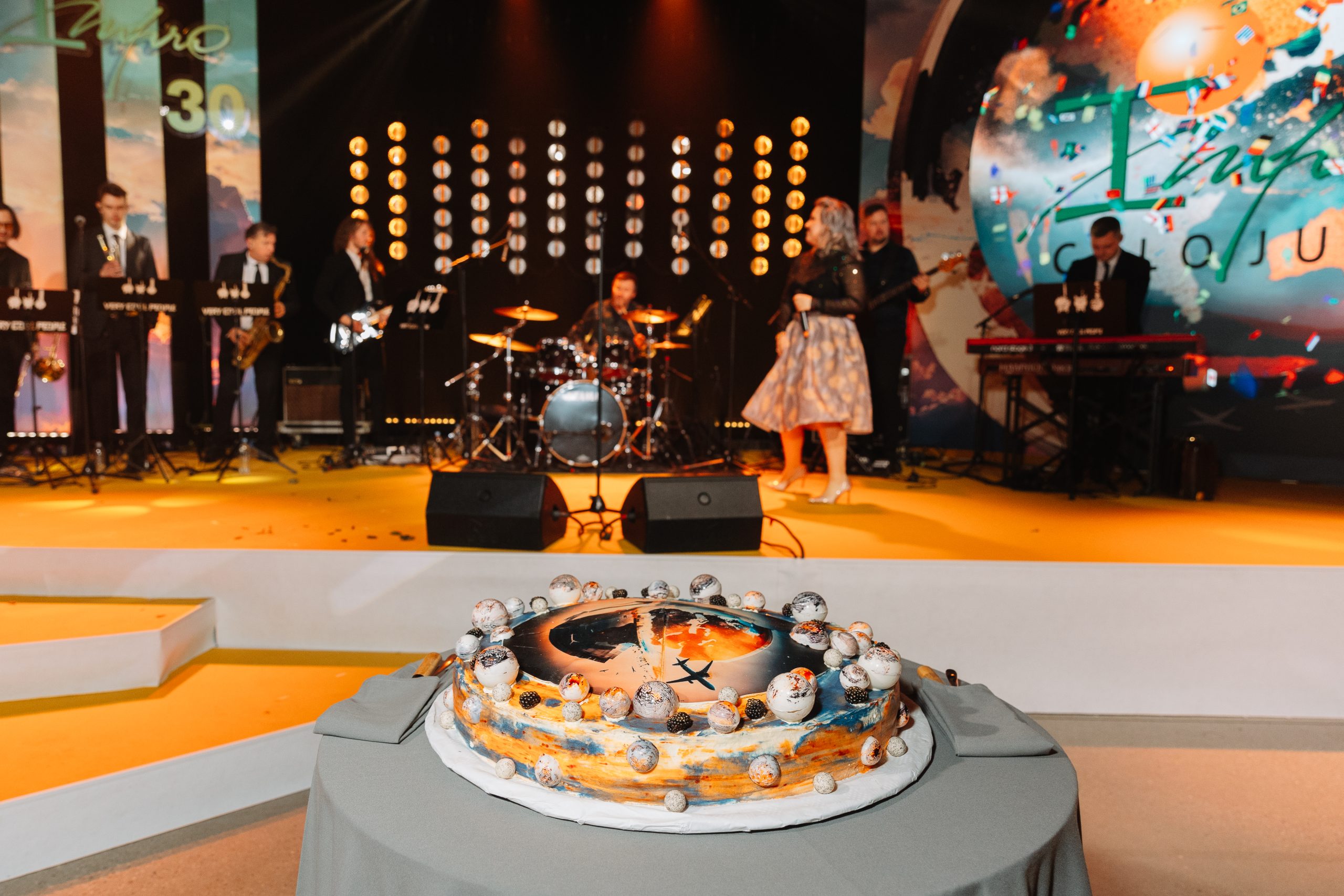 Svētku torte ar kosmosa tematikas dekoru priekšplānā, fonā muzikālā grupa uz skatuves jubilejas pasākumā.