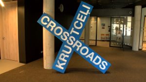 Liela zila izstādes instalācija ar uzrakstu "CROSSROADS KRUSTCELI" modernā iekštelpu vidē.